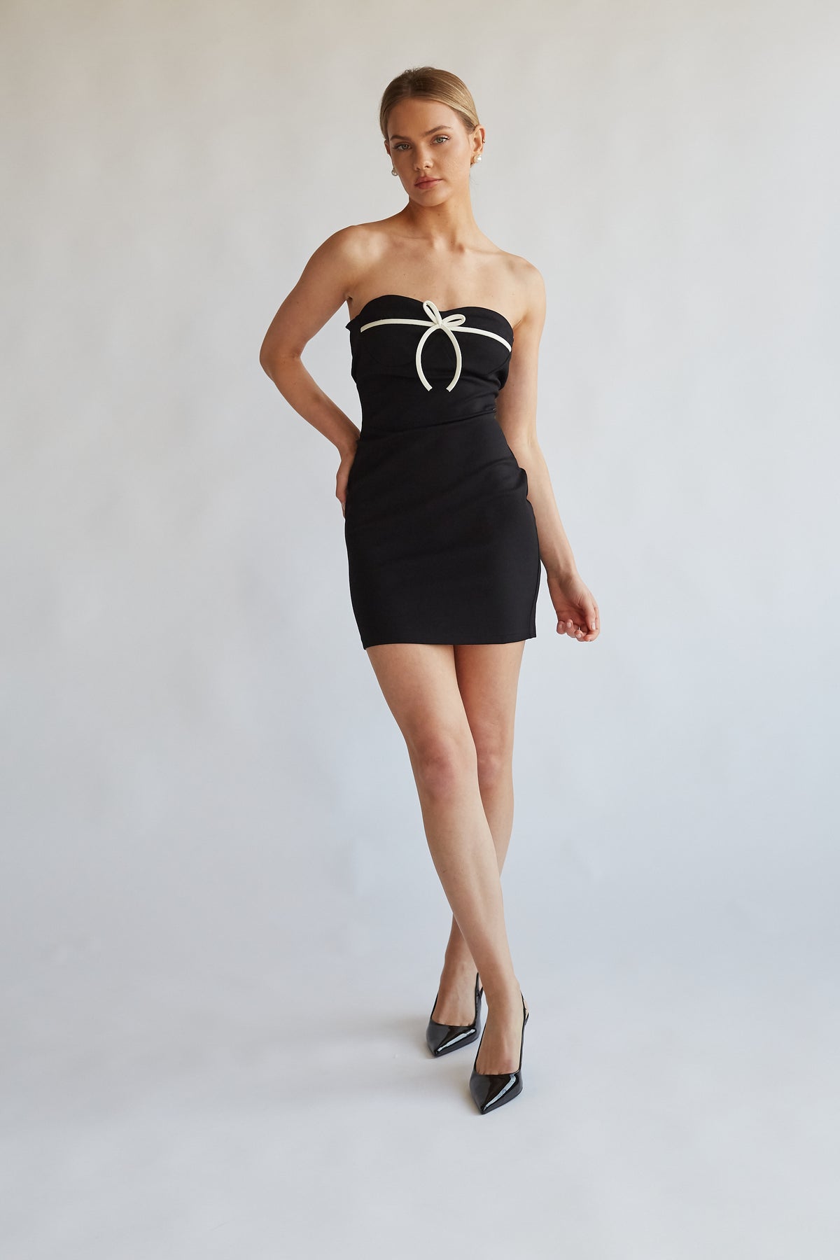 full body image of black mini dress with contrast white bow across bust | styled with pointed toe patent black pumps