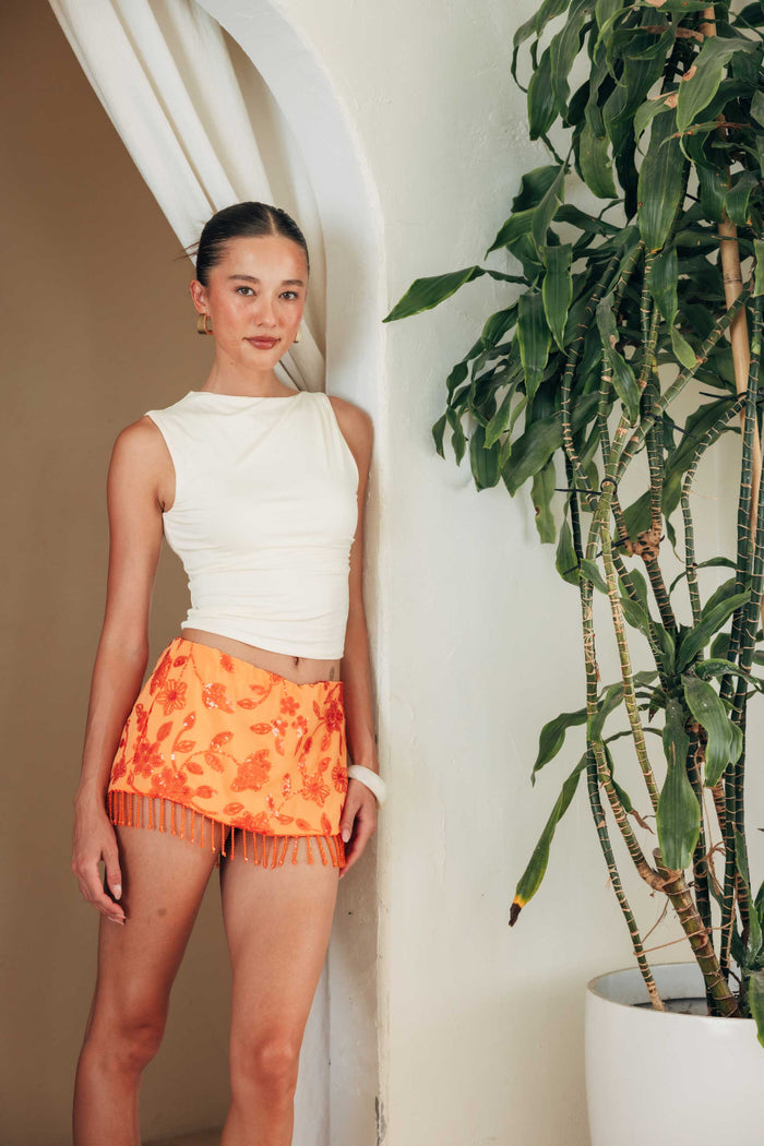 Woman wearing a white sleeveless top and orange floral skirt standing next to a plant.