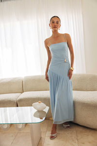Woman in a light blue dress standing in a living room with a beige sofa and glass coffee table.