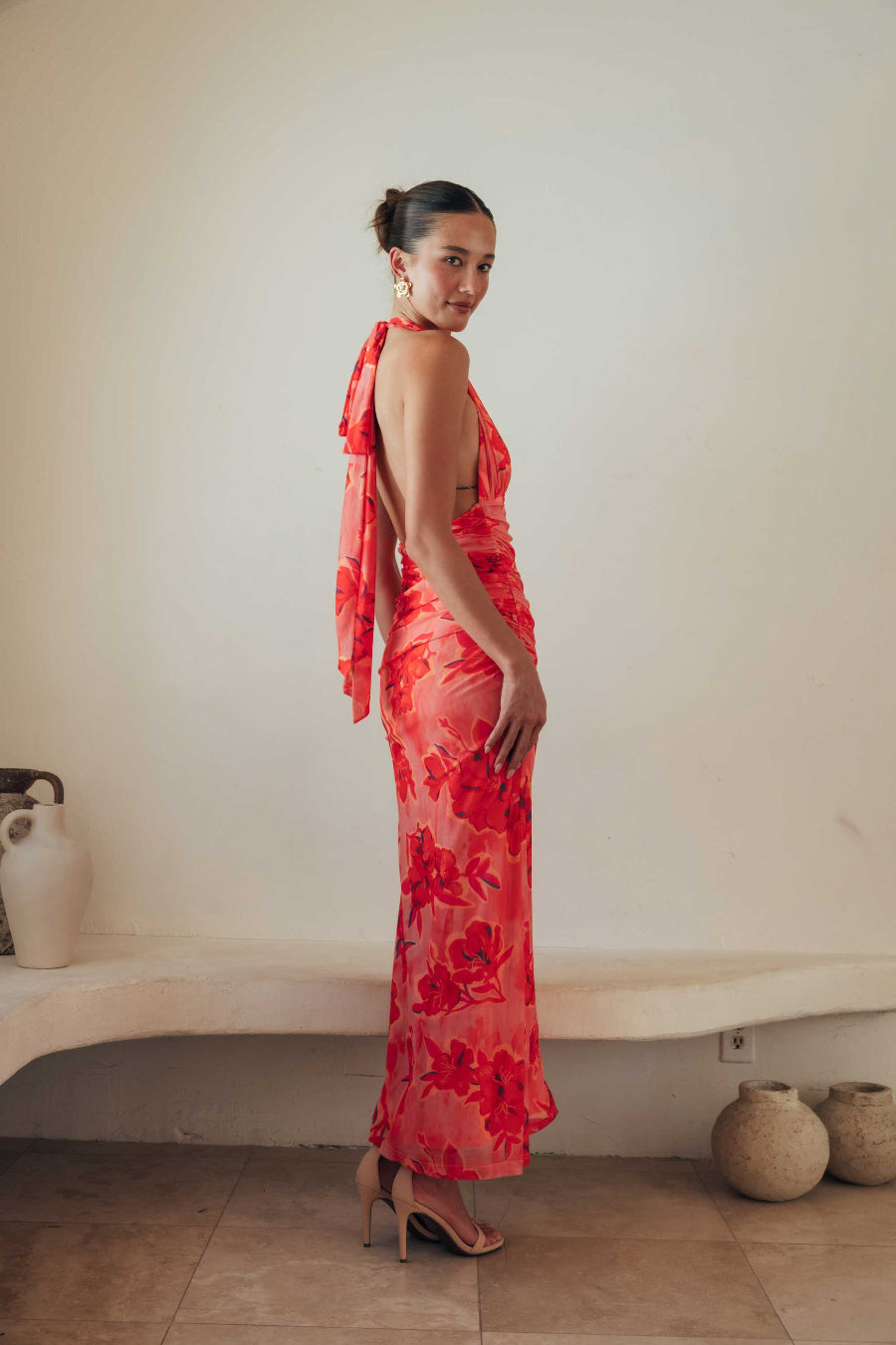 Woman in a red floral dress standing against a plain wall with decorative vases on a shelf.