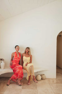 Two women sitting on a bench in a minimalistic setting with a white wall and tiled floor.