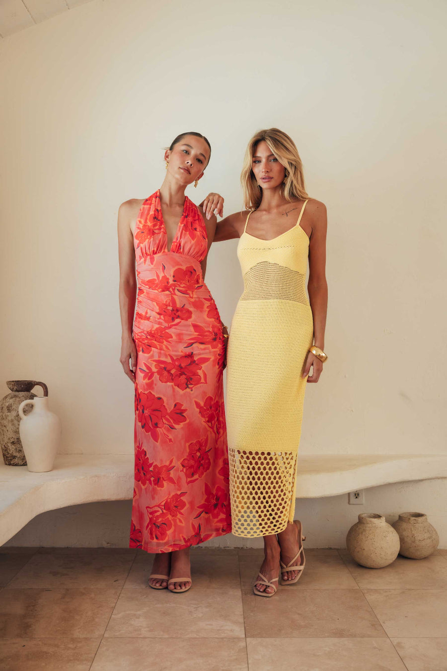 Two women standing side by side wearing colorful dresses in a minimal indoor setting.