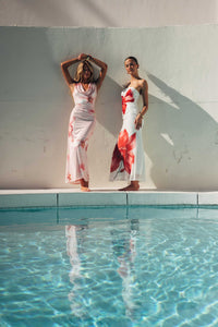 Two women in floral dresses standing by a pool with a white wall in the background.