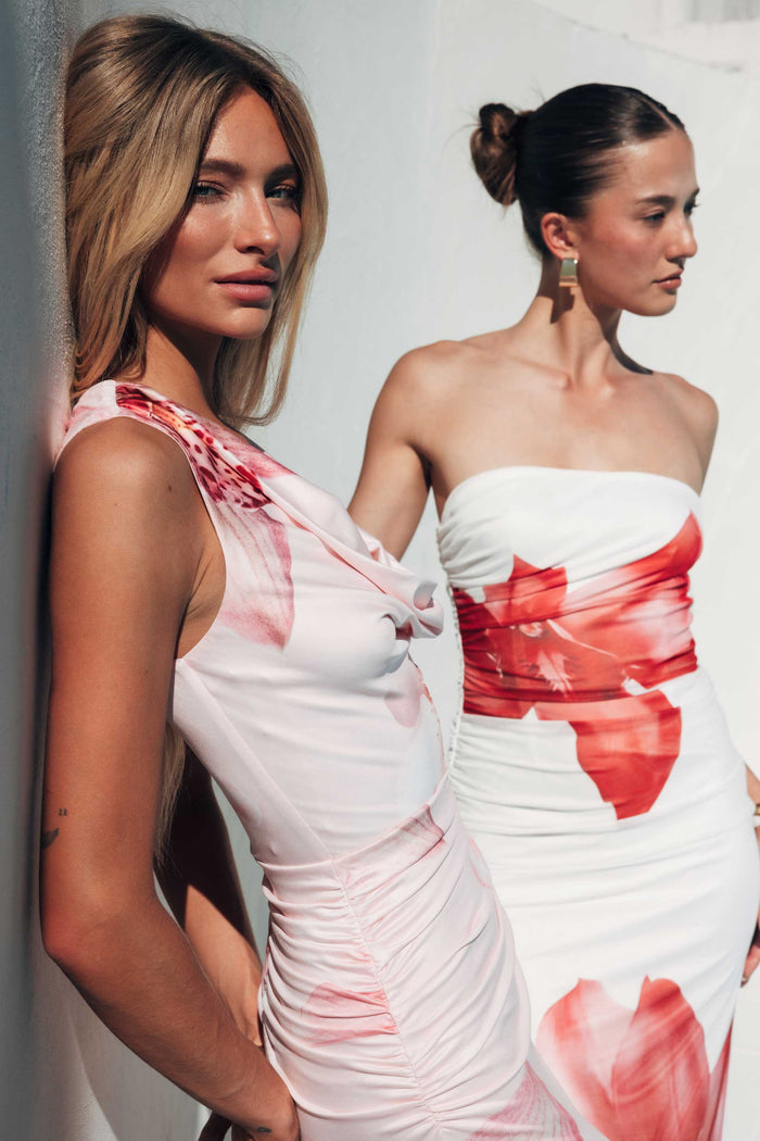 Two women wearing white dresses with red floral patterns against a plain background
