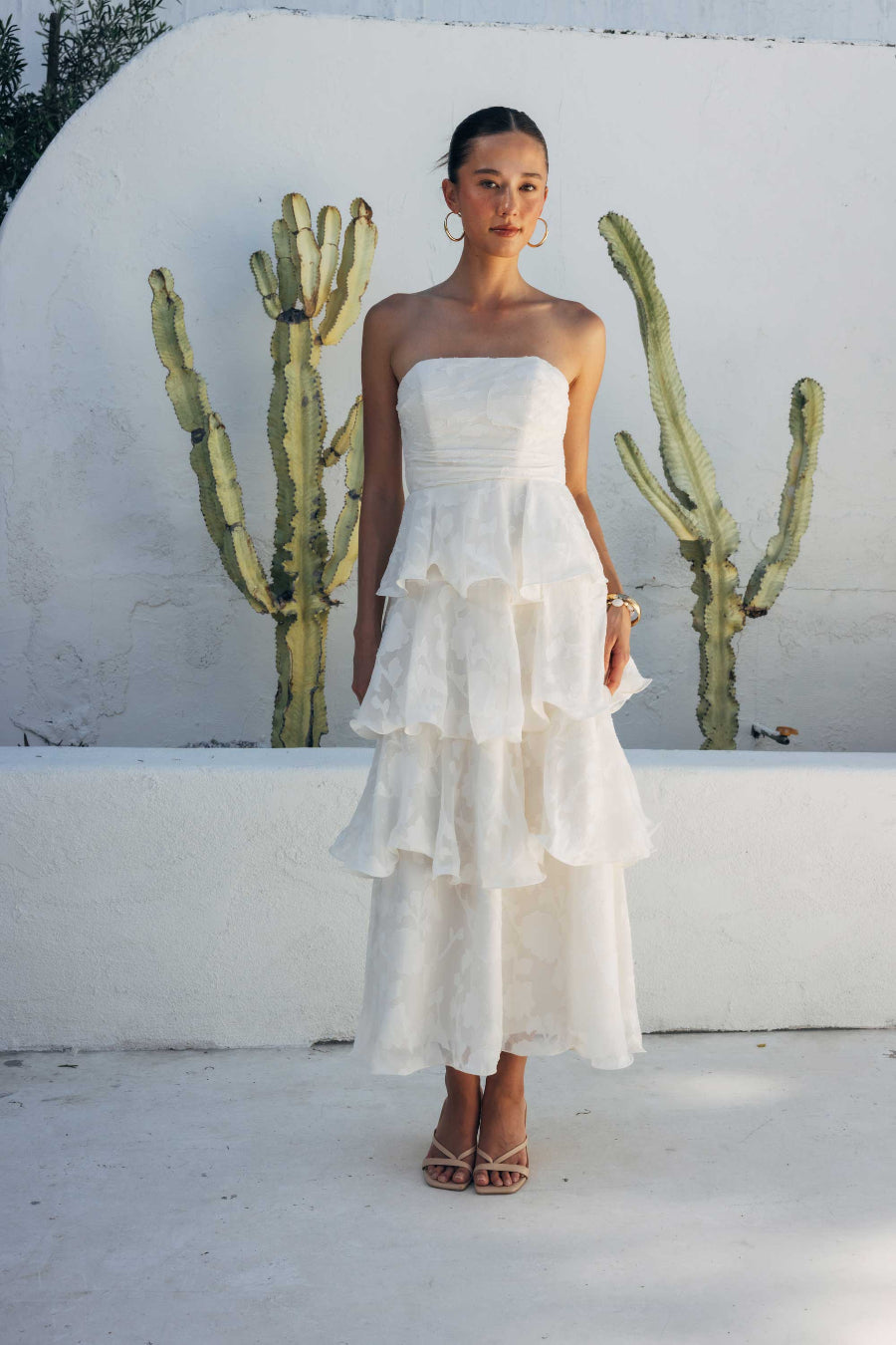 Woman in a white strapless dress standing in front of cacti against a white wall.