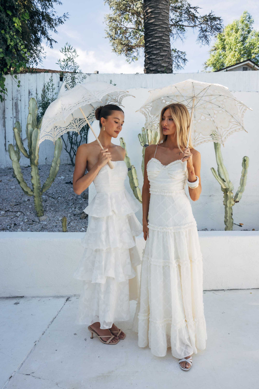 Two women in white dresses holding lace umbrellas against a white wall with cacti.