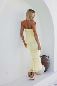 Woman in a yellow strapless dress standing against a white wall with a vase in the background.