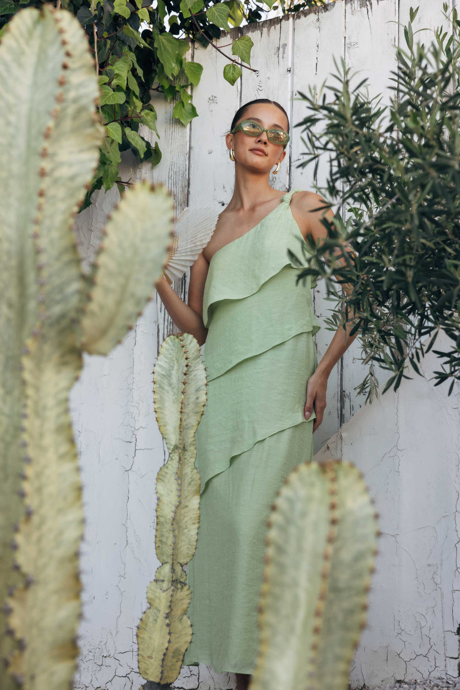 Woman in a green dress standing among cacti and plants against a white wooden fence.