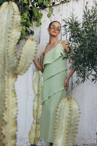 Woman in a green dress standing among cacti and plants against a white wooden fence.