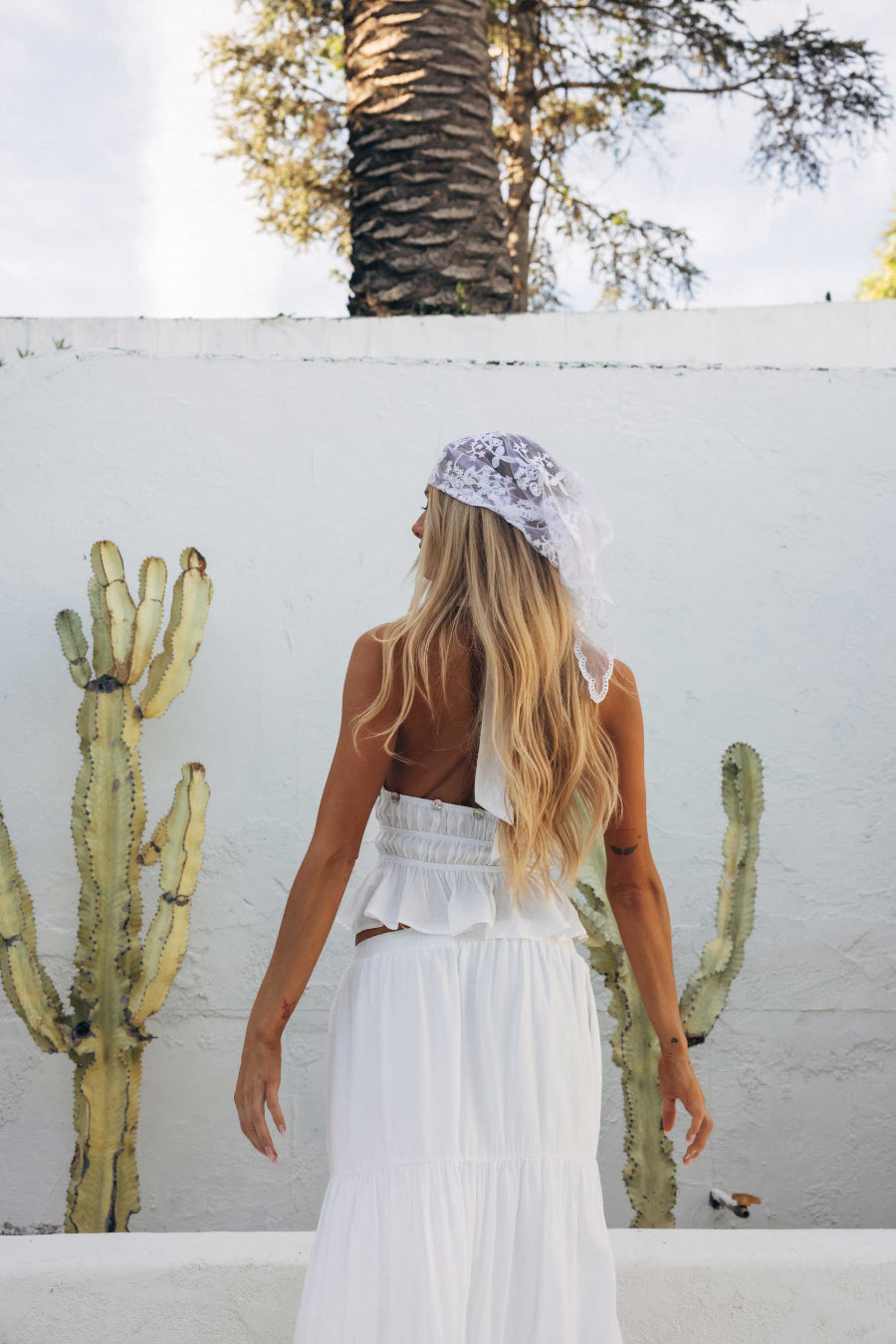 Woman in a white dress and headscarf standing against a white wall with cacti.