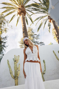 Woman in a white outfit standing in front of palm trees and a white wall.