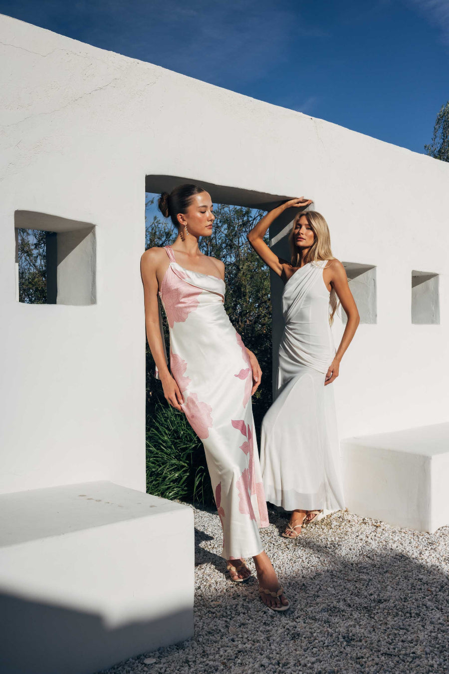 Two women in elegant dresses standing in front of a modern white building with blue sky.