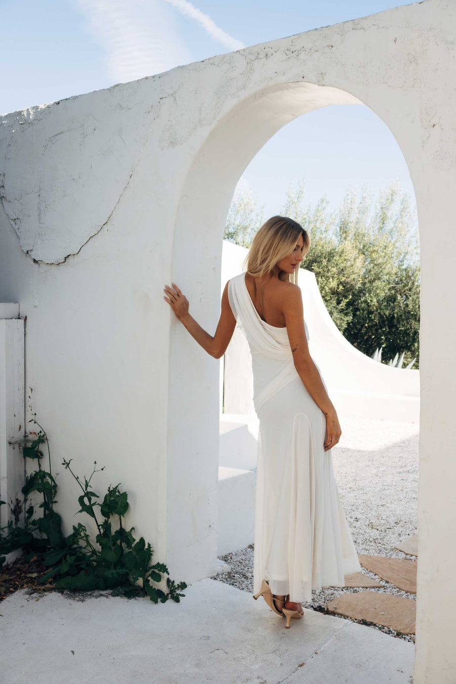 Woman in a white dress standing under a white archway with greenery in the background