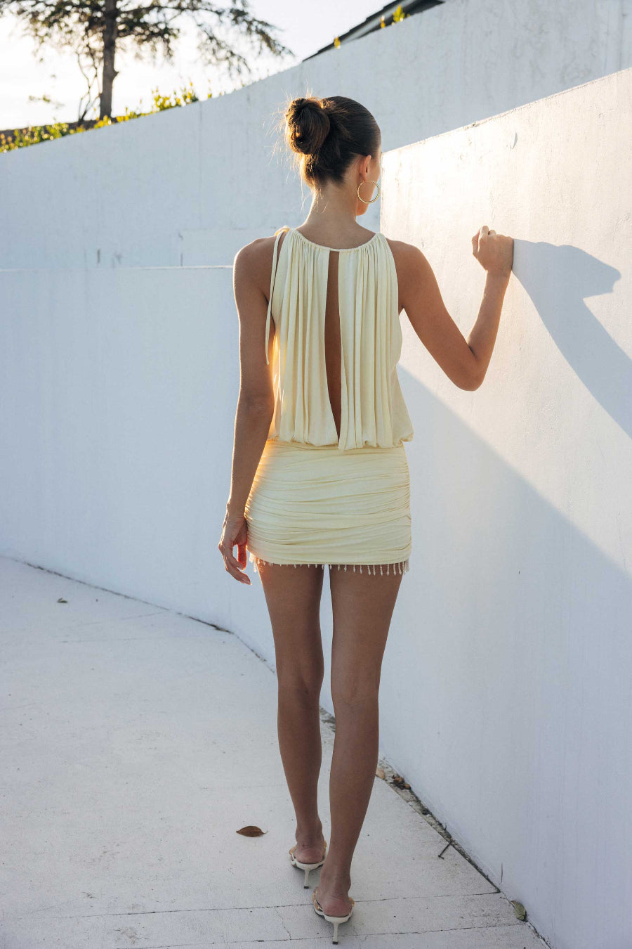 Woman in a yellow dress standing against a white wall with sunlight casting shadows.