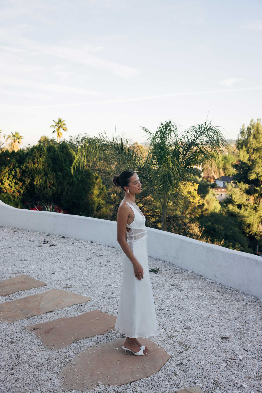 Woman in a white dress standing on a rooftop with greenery in the background