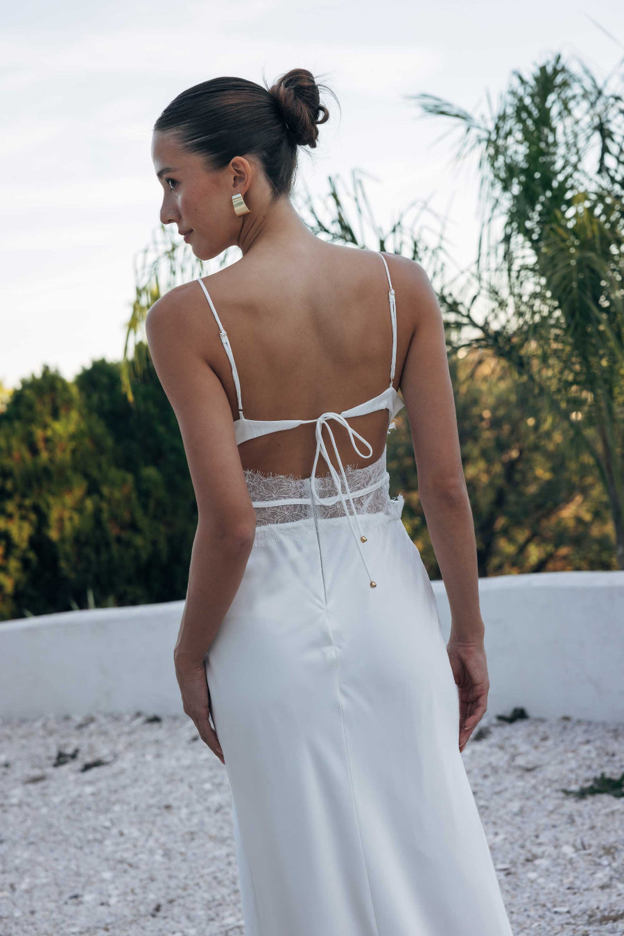 Woman wearing a white dress with an open back, standing outdoors with greenery in the background.