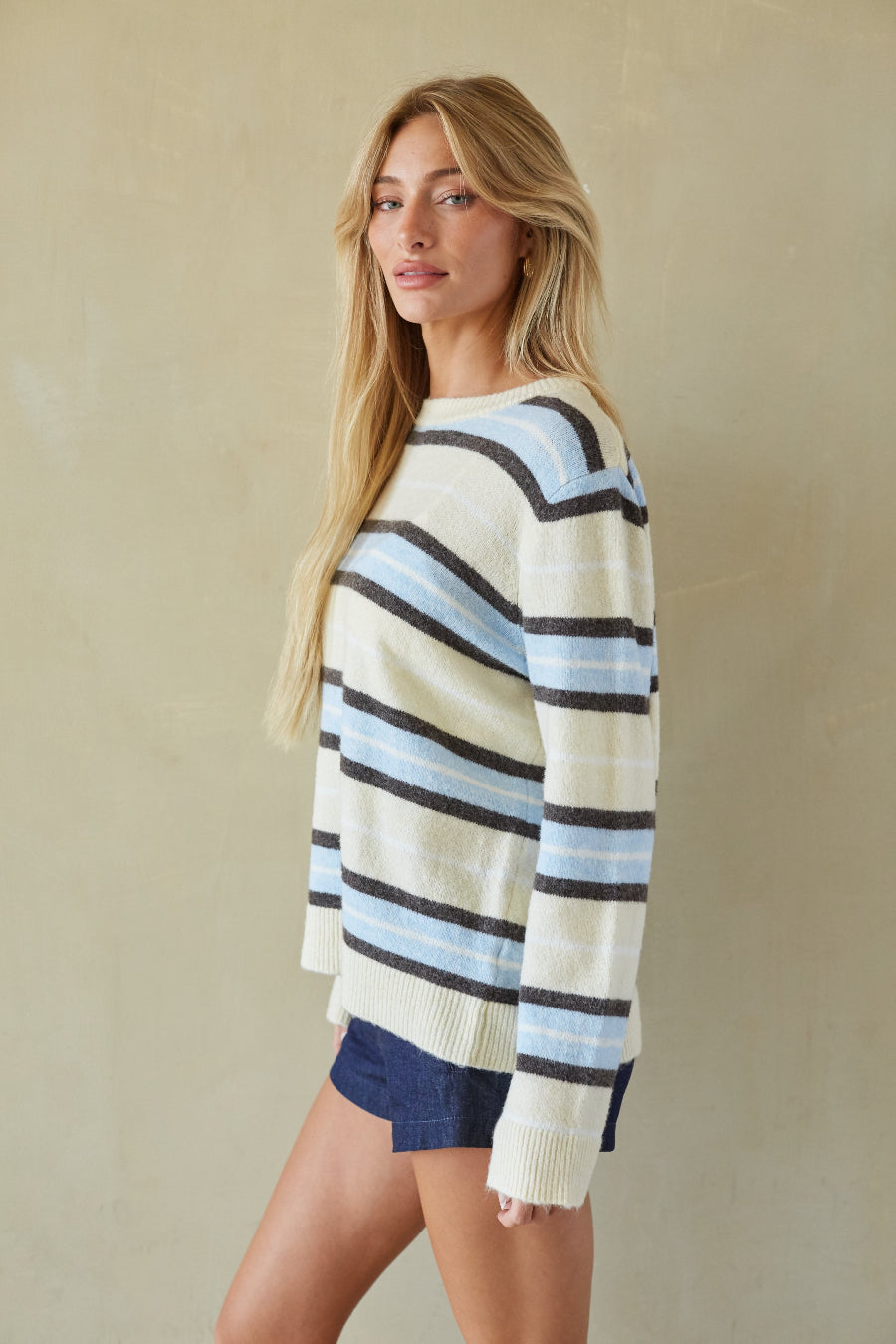 A woman wearing a cream knit sweater with horizontal stripes in light blue, navy, and black, paired with dark denim shorts, standing against a textured beige wall.