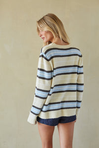 A woman wearing a cream knit sweater with horizontal navy, light blue, and dark charcoal stripes, paired with navy shorts, standing against a textured beige wall.