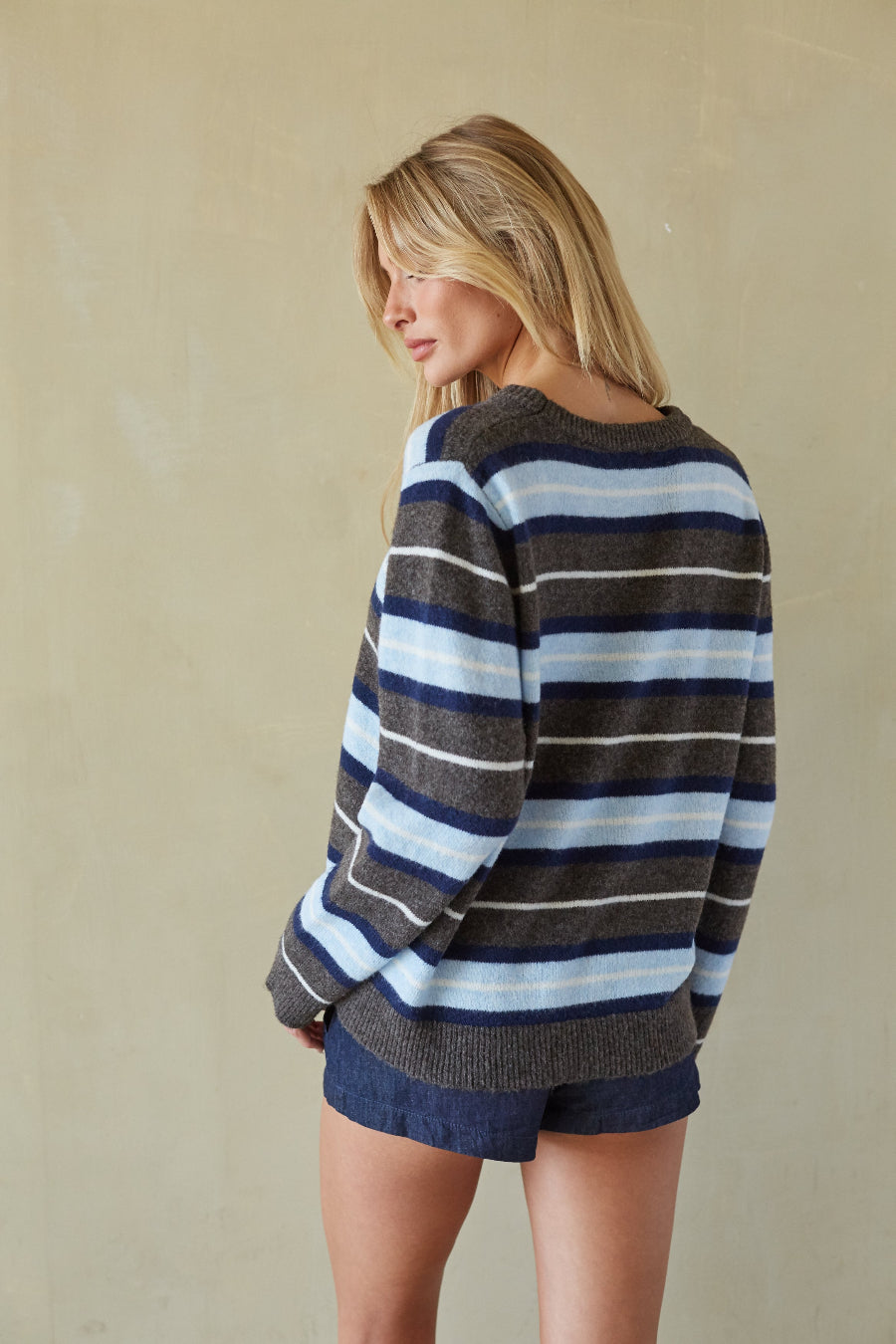 A woman wearing a striped knit sweater and denim shorts, photographed from the back against a plain beige wall.