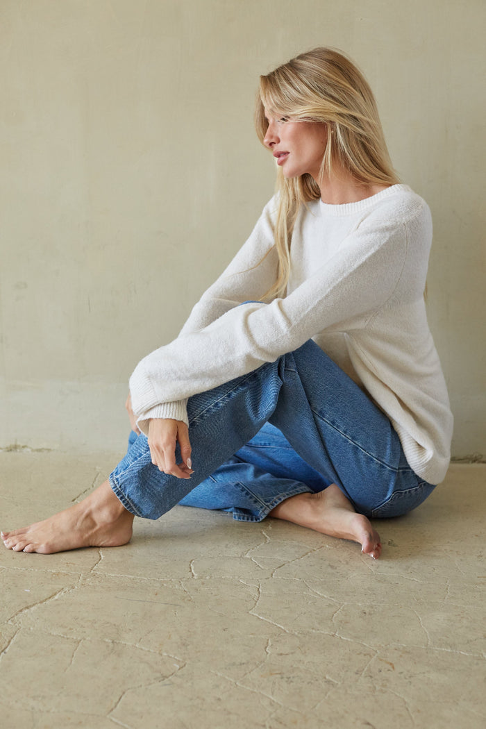 A woman sits barefoot on a textured beige floor against a matching beige wall, wearing a fuzzy off-white long-sleeve sweater and blue jeans.