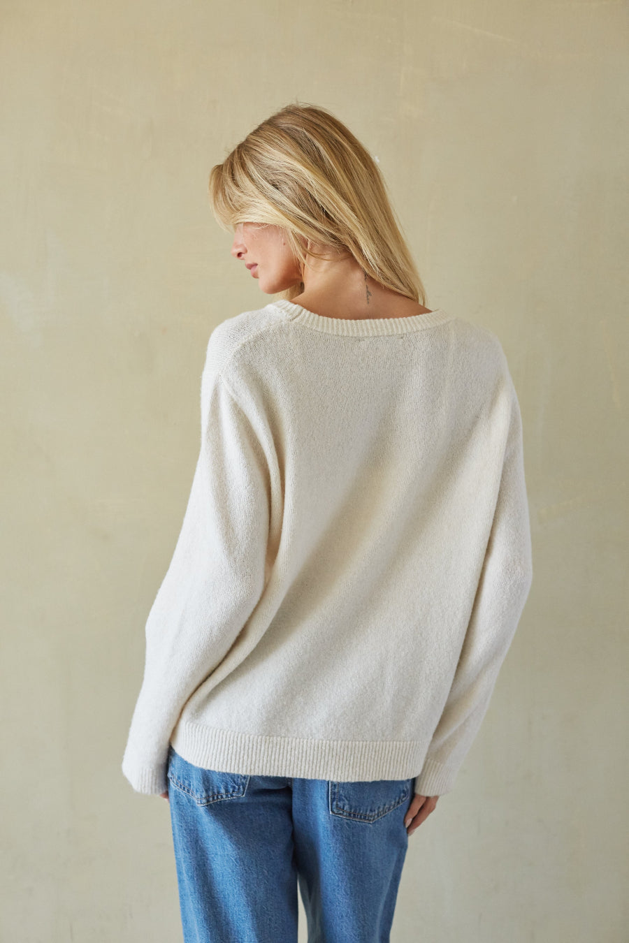 A woman with blonde hair is shown from behind wearing a cream-colored knit sweater and blue jeans, standing against a textured beige wall.