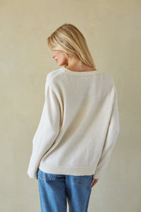 A woman with blonde hair is shown from behind wearing a cream-colored knit sweater and blue jeans, standing against a textured beige wall.