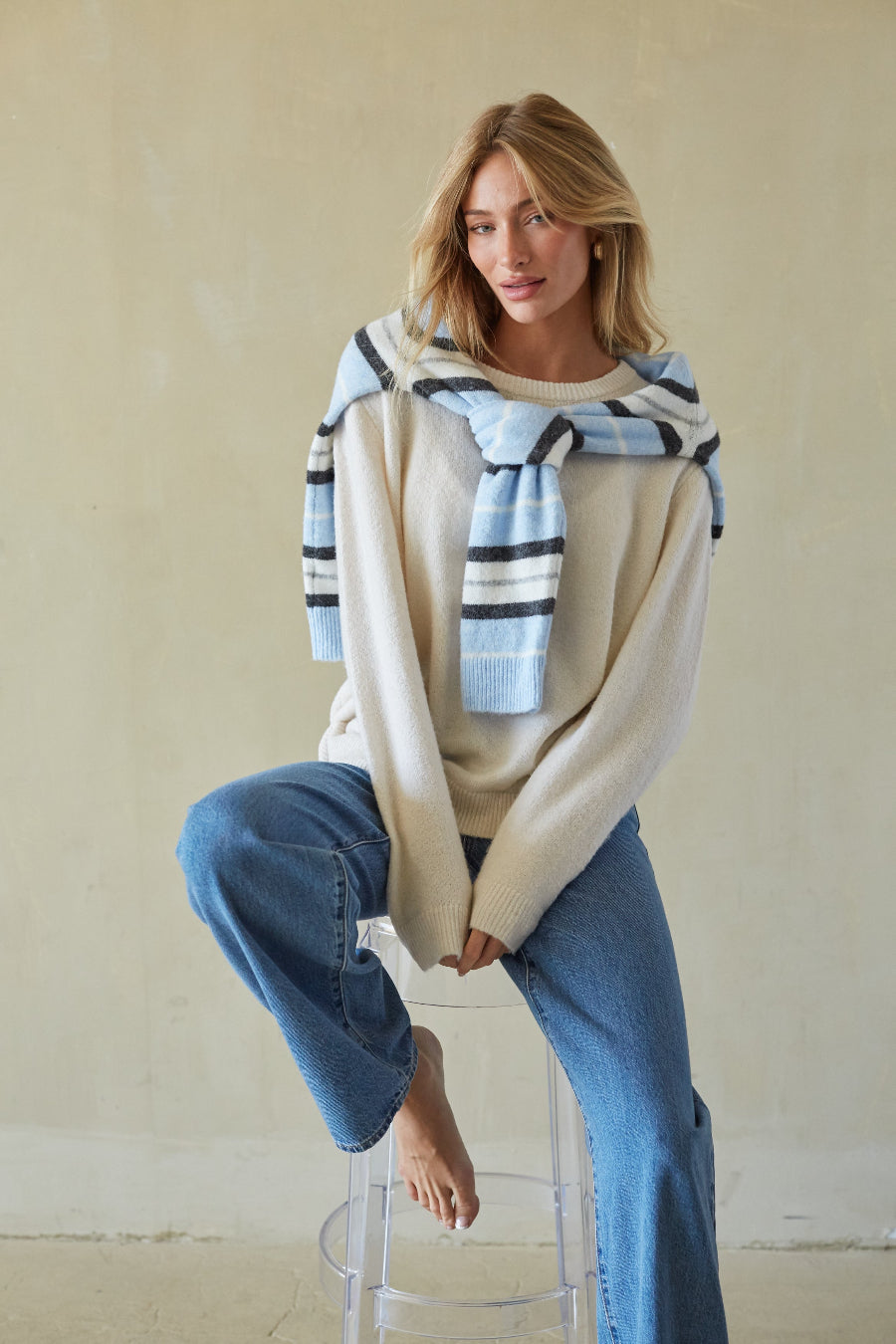 A woman sits barefoot on a clear stool against a textured beige wall, wearing a cream knit sweater with a light blue and navy striped knit scarf draped over her shoulders and blue jeans.