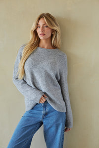 A woman with long blonde hair wearing a light gray knit sweater and blue jeans, posing against a textured beige wall.