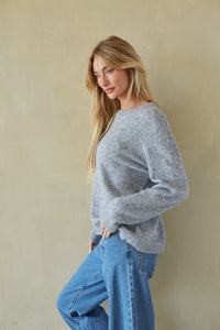 A woman with long blonde hair wearing a light gray knit crewneck sweater and blue jeans, standing against a textured beige wall.