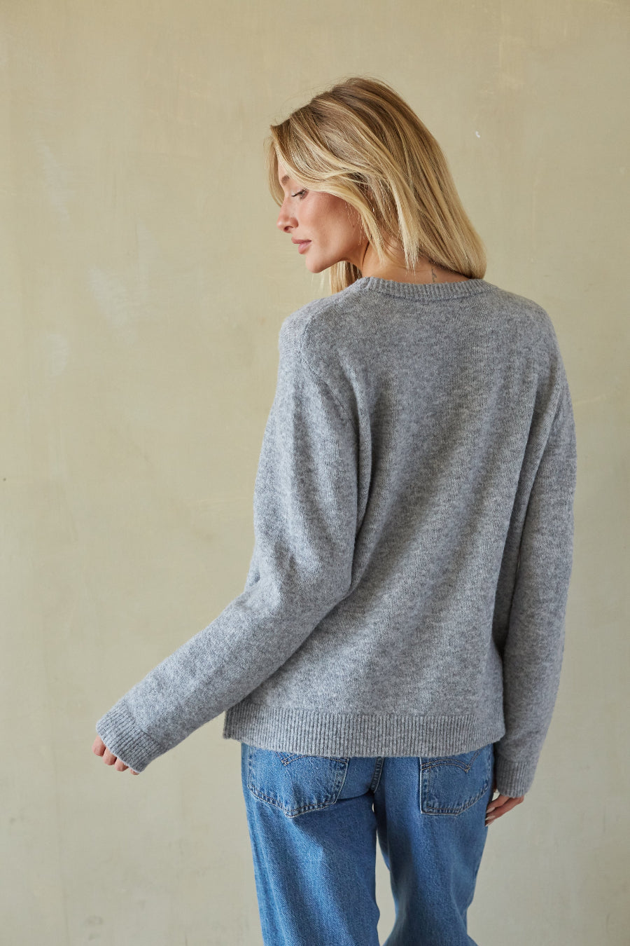A woman with blonde hair is shown from the back wearing a heather gray knit sweater and blue jeans, standing against a textured beige wall.