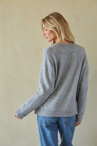A woman with blonde hair is shown from the back wearing a heather gray knit sweater and blue jeans, standing against a textured beige wall.