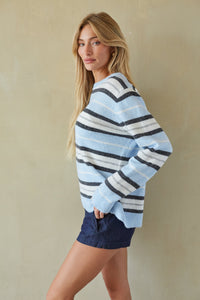 A woman modeling a light blue, navy, and white striped knit sweater, paired with dark blue shorts, against a textured beige wall.