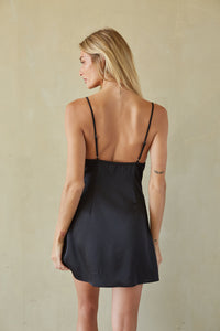 A woman shown from the back wearing a black spaghetti-strap slip dress against a textured beige wall.
