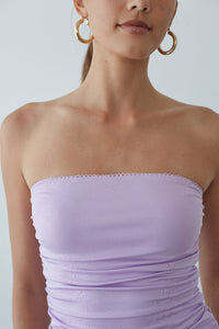 Woman wearing a strapless lavender dress with gold hoop earrings against a plain background