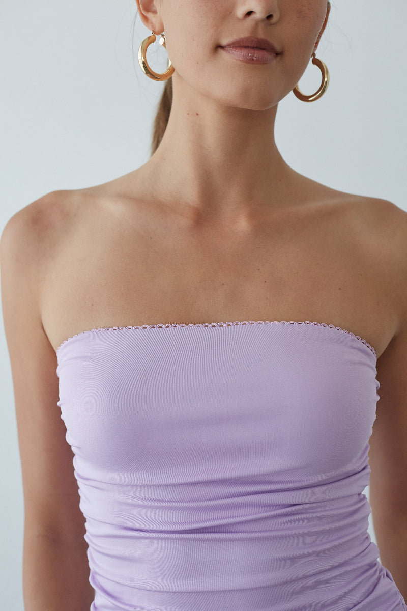 Woman wearing a strapless lavender dress with gold hoop earrings against a plain background