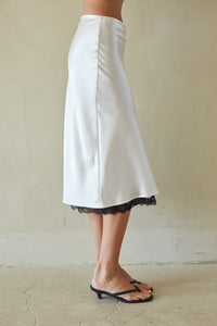 A person wearing a white midi skirt with a black lace hem, paired with black heeled thong sandals, standing against a textured beige wall.