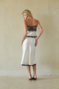 A woman stands with her back turned wearing a white satin slip dress with black lace trim on the straps, bodice, and hem, paired with black heels, against a textured beige wall.