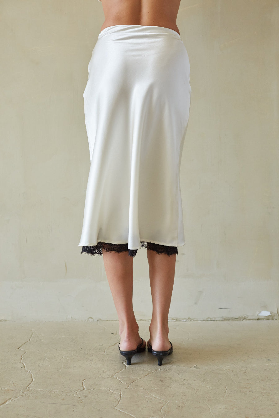 A person shown from the back wearing a white satin midi skirt with black lace trim and black pointed-toe mule heels, standing against a plain beige wall and concrete floor.