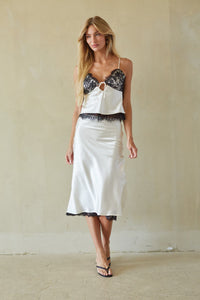 A woman modeling a white satin slip dress with black lace trim and a matching white camisole top, standing against a textured beige wall.