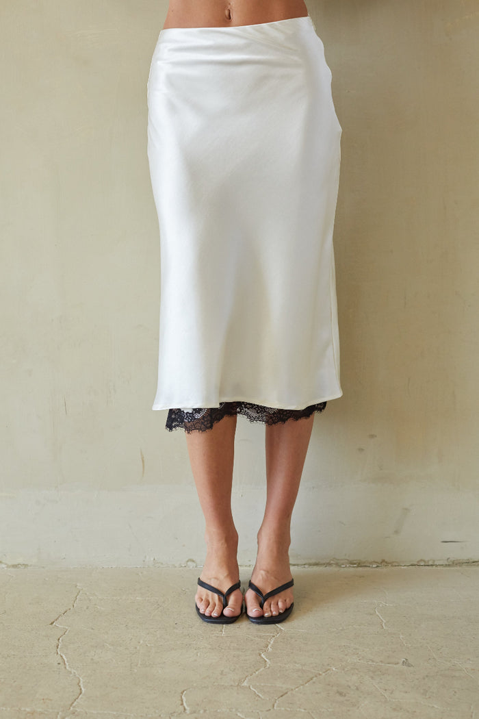 A white satin midi skirt with a black lace hem, worn by a model standing against a textured beige wall.