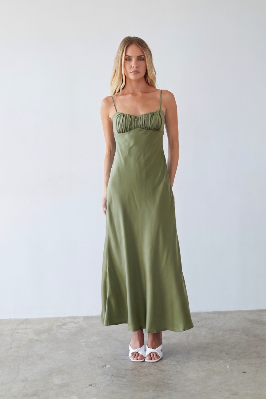 green-image | A woman modeling a long olive green satin maxi dress with thin spaghetti straps and a ruched sweetheart neckline, standing against a neutral light gray wall and concrete floor.