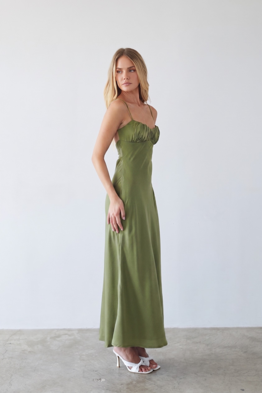 A woman modeling a long olive green satin maxi dress with thin spaghetti straps and a gathered sweetheart neckline, standing against a minimalist white wall and concrete floor.