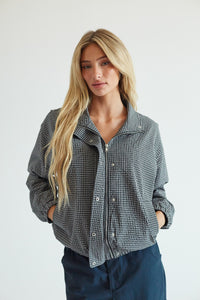 A blonde woman wearing a gray and black gingham zip-up shirt jacket with dark blue pants, standing against a plain light gray background.
