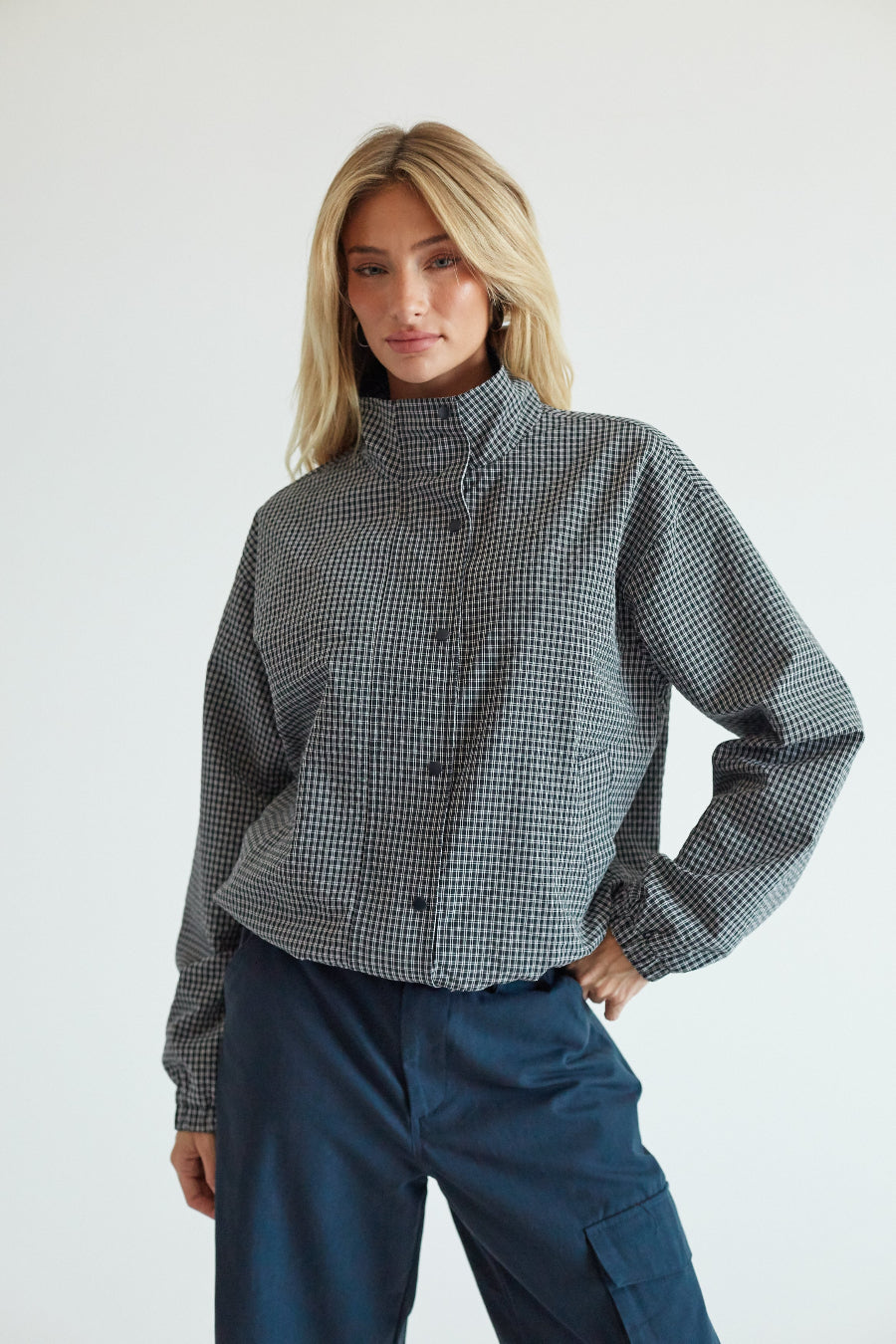 A woman modeling a cropped, long-sleeve button-up shirt with a black and white micro-check pattern and a high mock neck, paired with dark blue cargo pants against a plain light gray background.