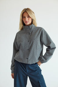 A woman modeling a cropped, long-sleeve button-up shirt with a black and white micro-check pattern and a high mock neck, paired with dark blue cargo pants against a plain light gray background.