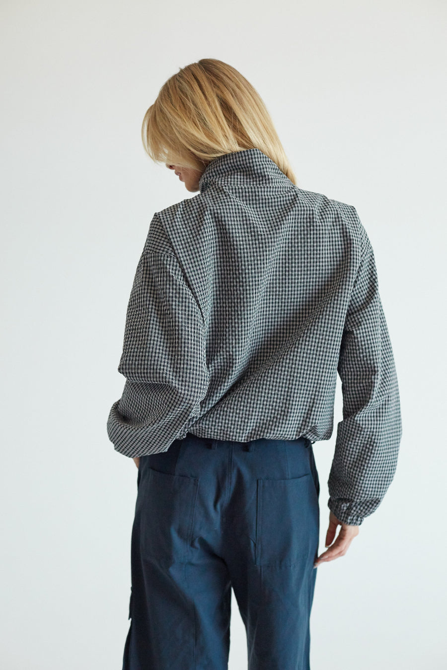 A model viewed from behind wearing a black and white micro-check patterned mock-neck shirt and dark blue trousers, set against a plain light gray background.