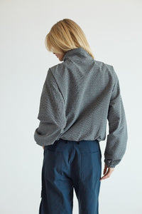 A model viewed from behind wearing a black and white micro-check patterned mock-neck shirt and dark blue trousers, set against a plain light gray background.