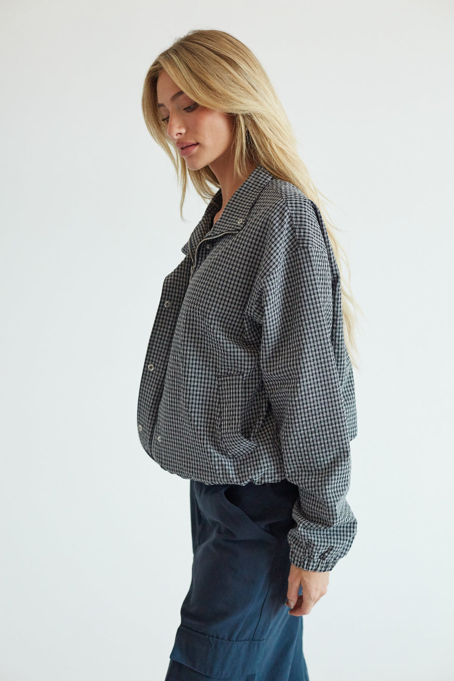 A woman wearing a black and white gingham cropped button-up shirt and dark blue pants, photographed against a clean white studio background.