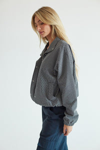 A woman wearing a black and white gingham cropped button-up shirt and dark blue pants, photographed against a clean white studio background.