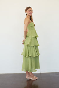 A woman modeling a light green tiered maxi dress with side cutouts, standing in profile against a plain white wall.
